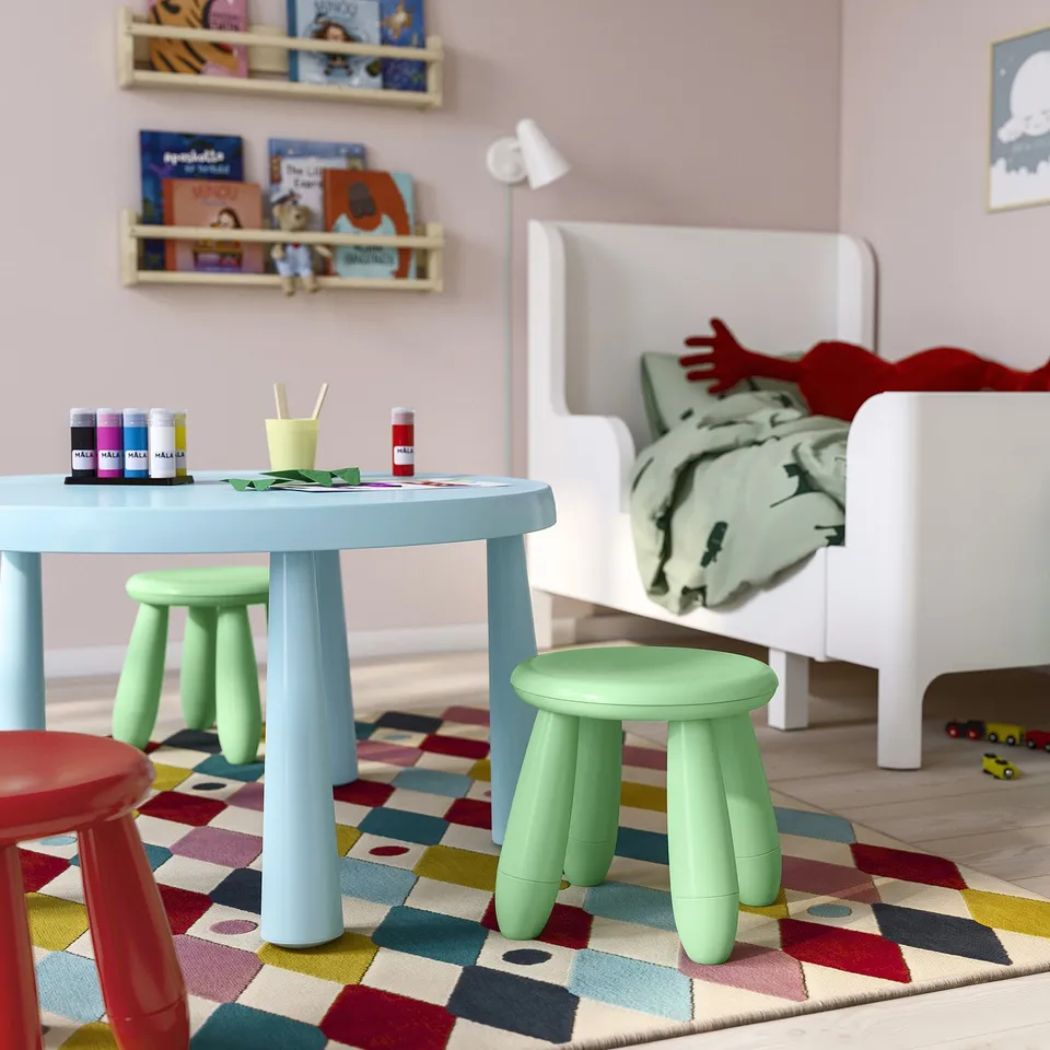Kids playroom with light blue round table, green stools, colourful rug. Table has craft supplies, bookshelves, white bed in background.