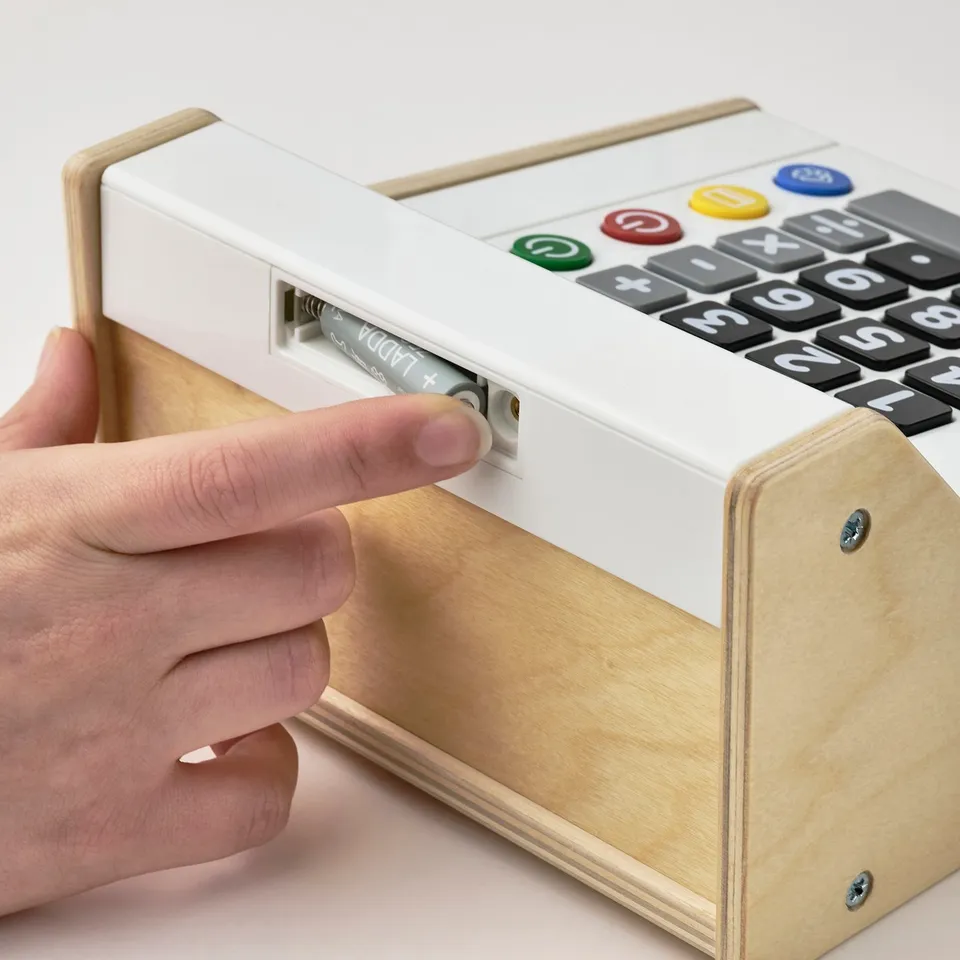 Person inserting battery into a toy cash register. The cash register has a calculator and play money slot.