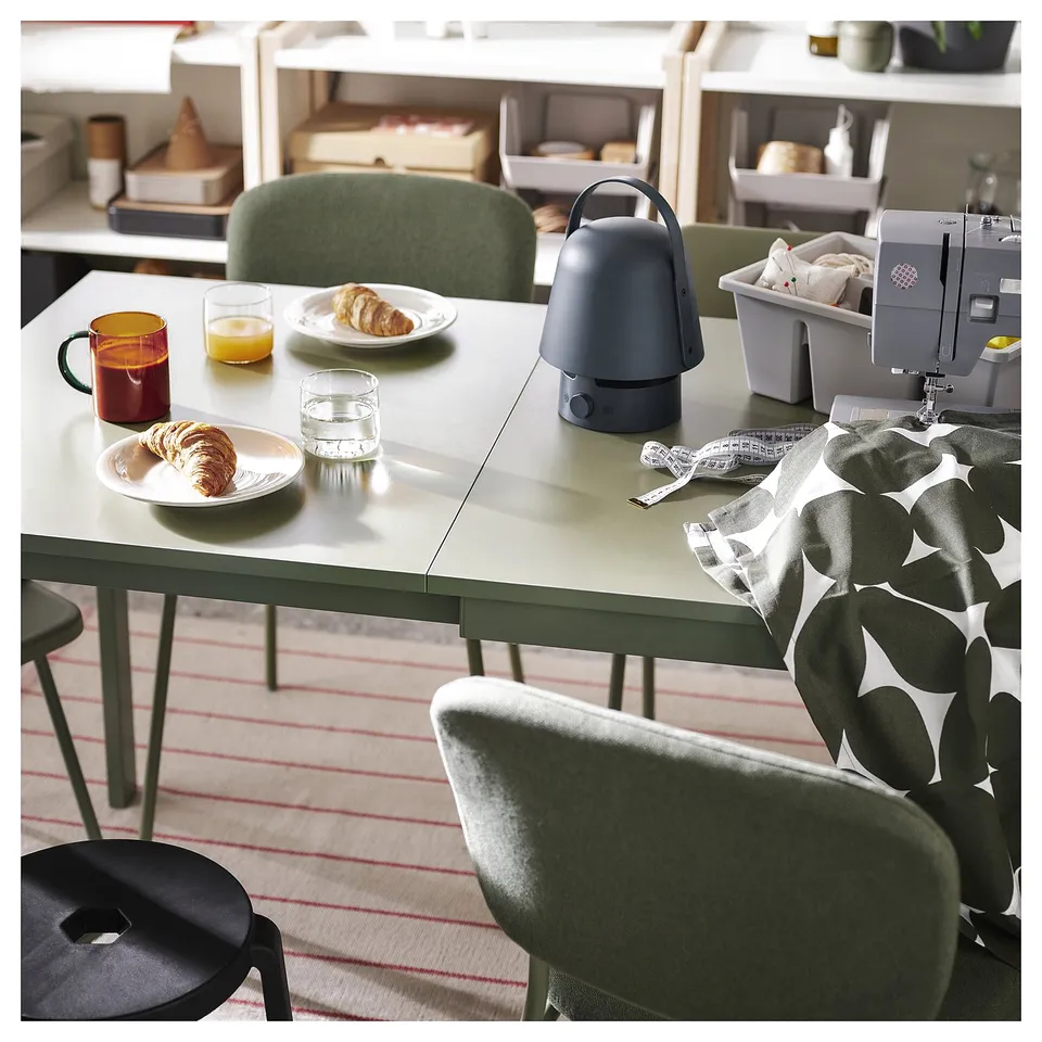 Cosy dining area with green table, chairs, and striped rug. Table has pastry, juice, mug, and lamp. Green stool and sewing machine.