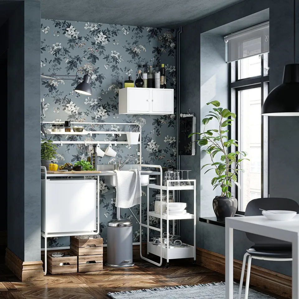 Modern kitchen with blue floral wallpaper, white furniture, including a small fridge, shelves with dishes, and a window with a plant.