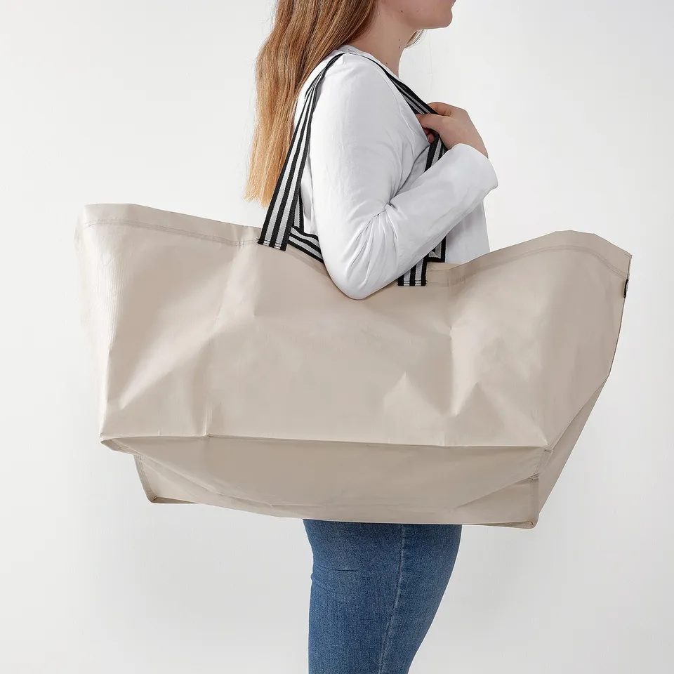 Person holding a large beige GÖRSNYGG tote bag with striped handles, designed for shopping, laundry, or travel.