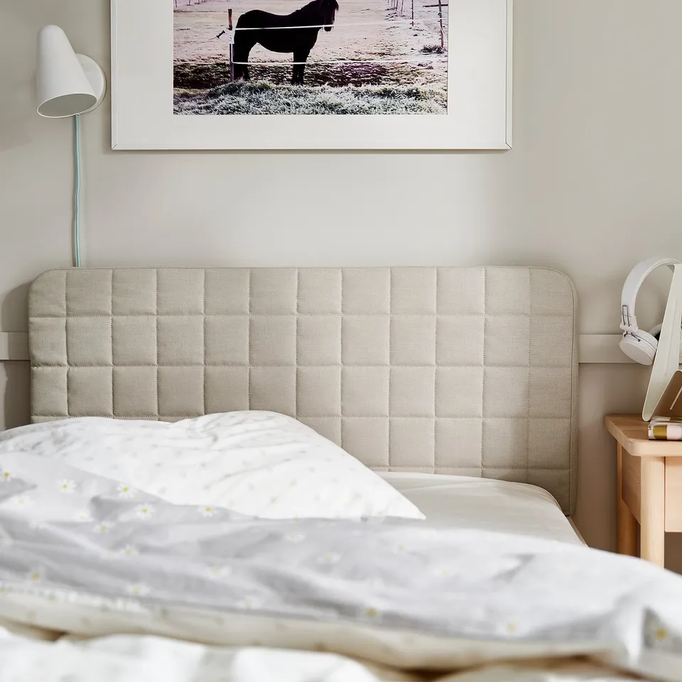 Minimalist bed with beige padded headboard, white bedding, and wall art. A bedside lamp and headphones on the wooden bedside table.