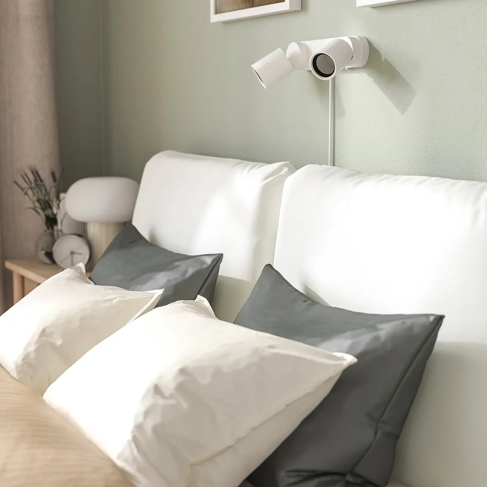 A minimalist bed with two pillows, featuring a gently sloped headboard, positioned against a green wall. A wall-mounted white lamp is above it.