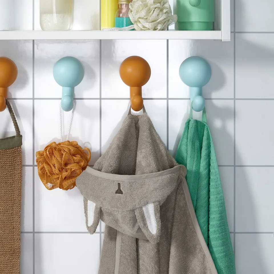 Three hooks on a tiled wall hang items: a grey bunny towel, orange loofah, and green cloth.