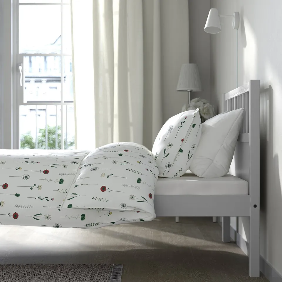 Bedroom with a white SMYGA bed by a window. The bed has floral bedding and is positioned beside a lamp and curtains.