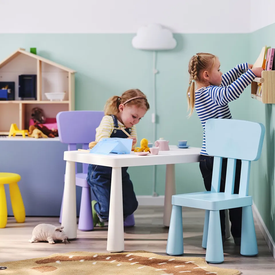 Two kids playing with toys at a MAMMUT table, one reaching for a toy on a shelf in a colourful room.