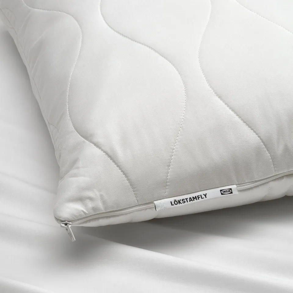 Close-up of a white ergonomic pillow, labelled LÖKSTAMFLY, showcasing its soft, quilted cover and zip detail.