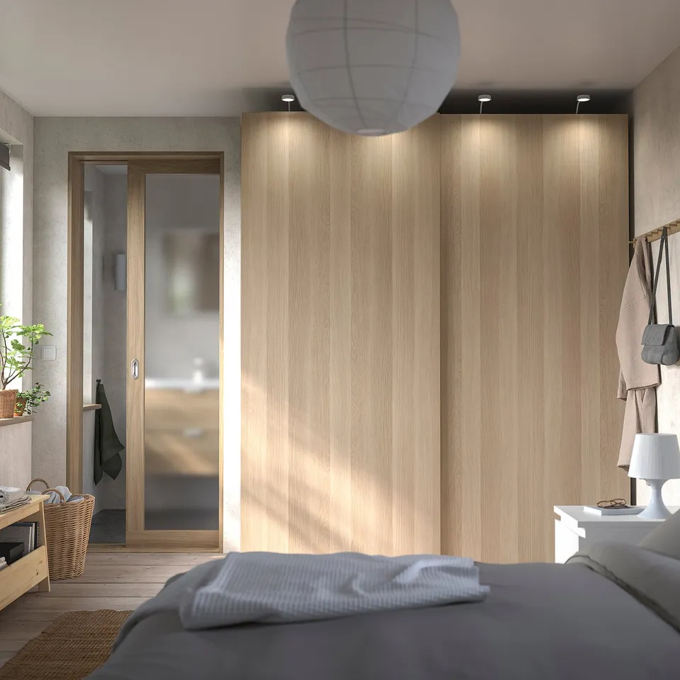 A modern bedroom with light wooden PAX wardrobes featuring sliding doors. Theres a grey bed with a white blanket.