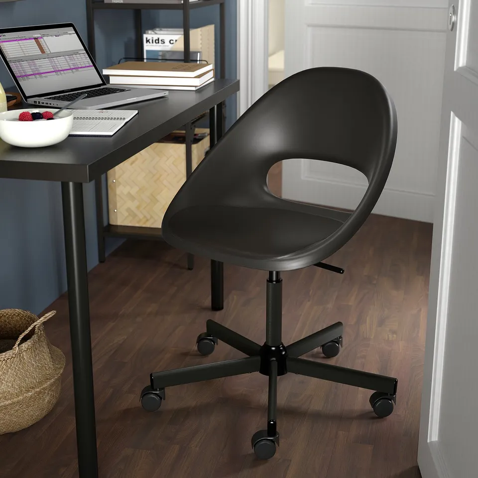 Black office chair with cutout back on dark wooden floor. Desk with laptop and bowl of berries.