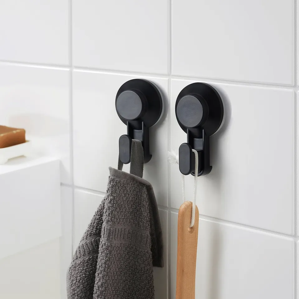 Two black TISKEN wall hooks hold a grey towel and wooden spatula. Small, round, suction-cup design, mounted on white tiled wall.