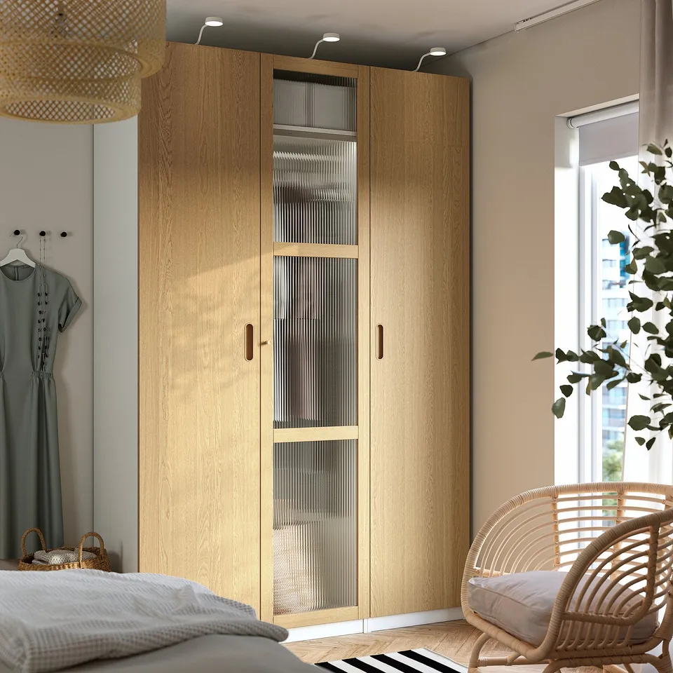 Brown PAX wardrobe with glass centre, clothes left, wicker chair by window, bed with grey blanket.