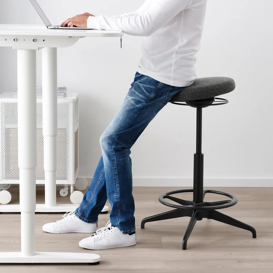 Person sits on adjustable stool at standing desk, promoting active posture and movement.