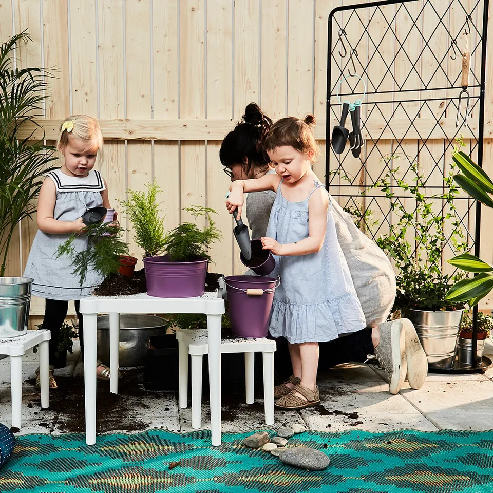 Two girls in light dresses garden outdoors, using tools. An adult assists, surrounded by plants.