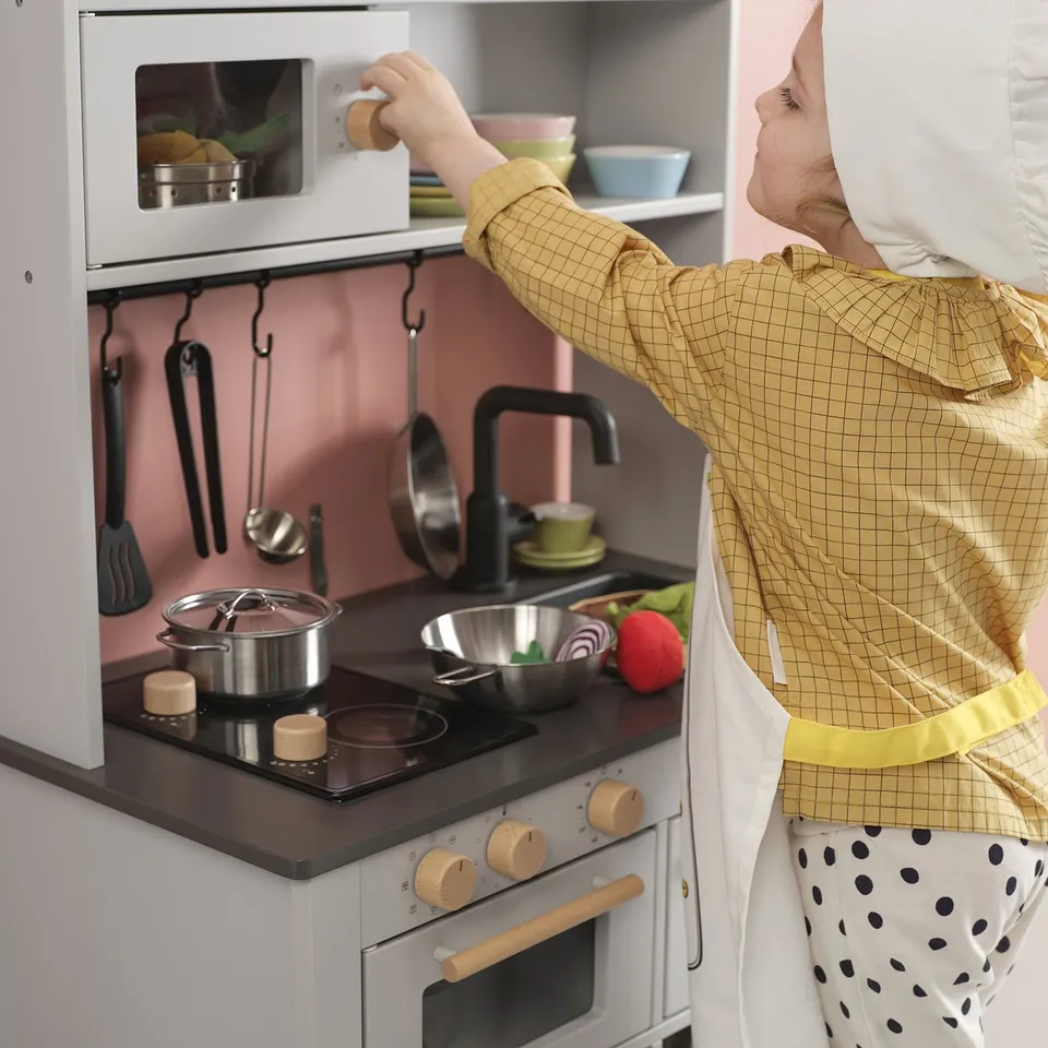 Child playing with toy kitchen set, including microwave, stove, sink, and utensils, learning cooking roles and safety.