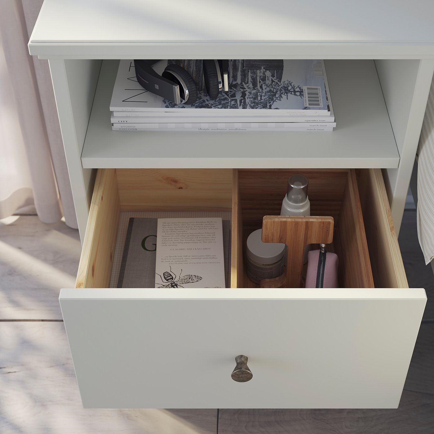 White IDANÄS bedside table, open drawer with small items, top shelf with books.