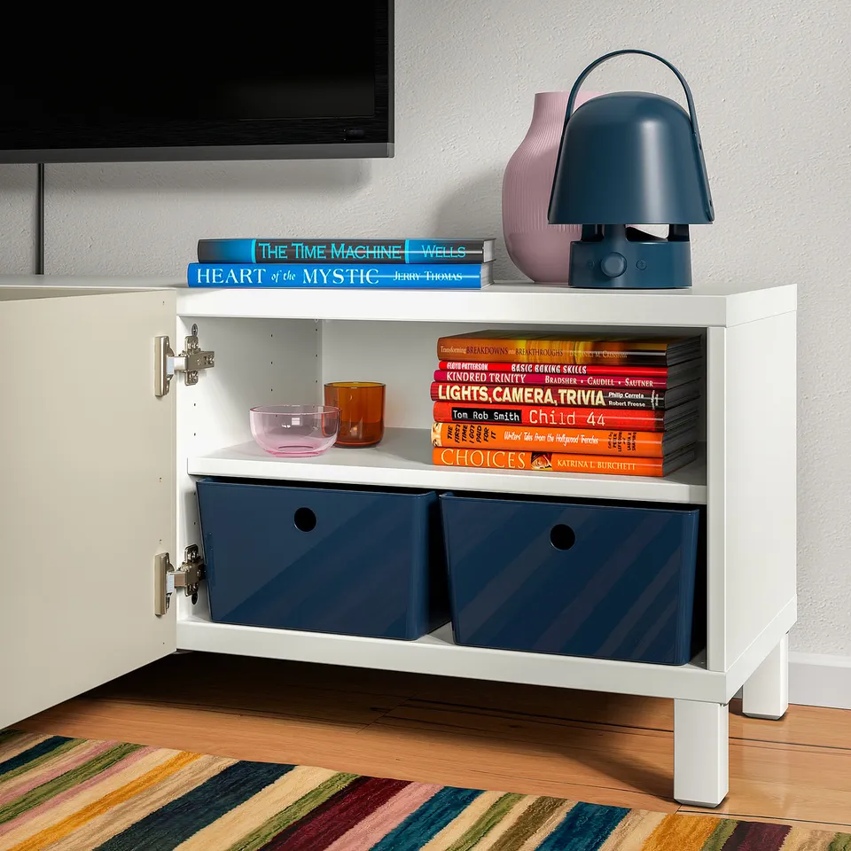 White storage cabinet with wooden floor, colourful rug, books, two blue storage bins, various decorative items, and a blue lamp on top.