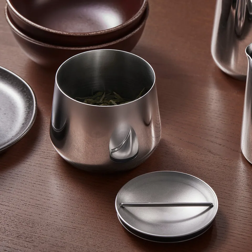 Metal jar with lid on wooden table. Contains green tea leaves, part of a stylish kitchen set.