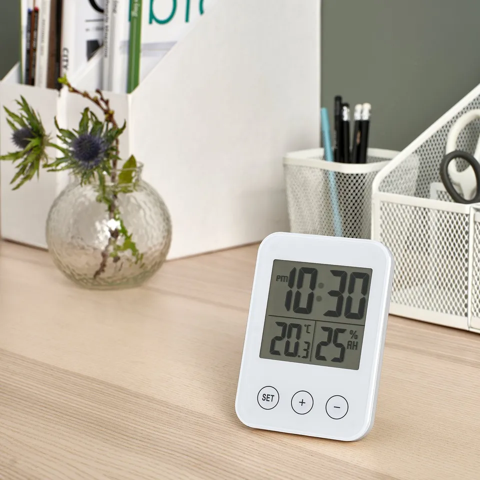 Desk scene with white SLÅTTIS multi-function clock showing time, temperature, humidity. Includes small decorative plant & pens. Clock is compact.