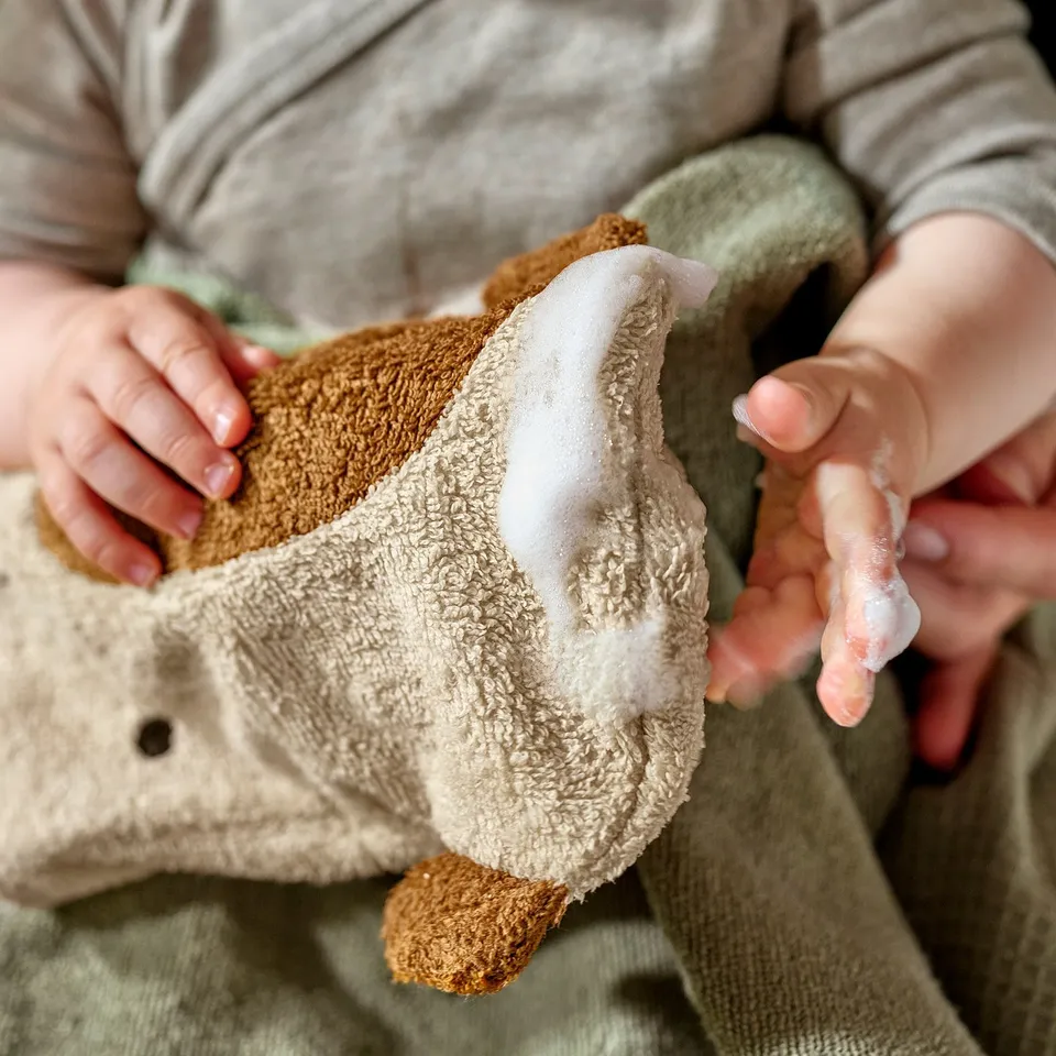 Baby holding beige GULDVÄVARE mitt. Soft, textured, embroidered face design. Perfect for gentle cleaning.