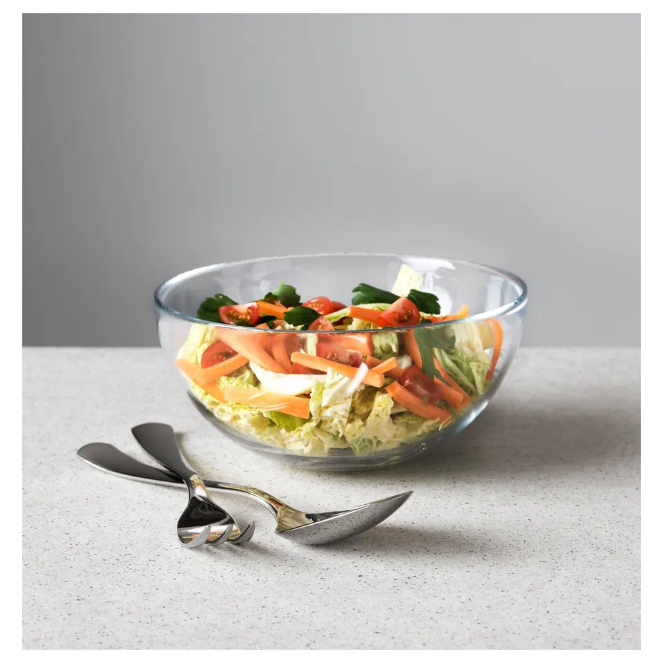 A clear glass bowl filled with salad sits on a speckled counter. Two mismatched serving spoons rest beside it.