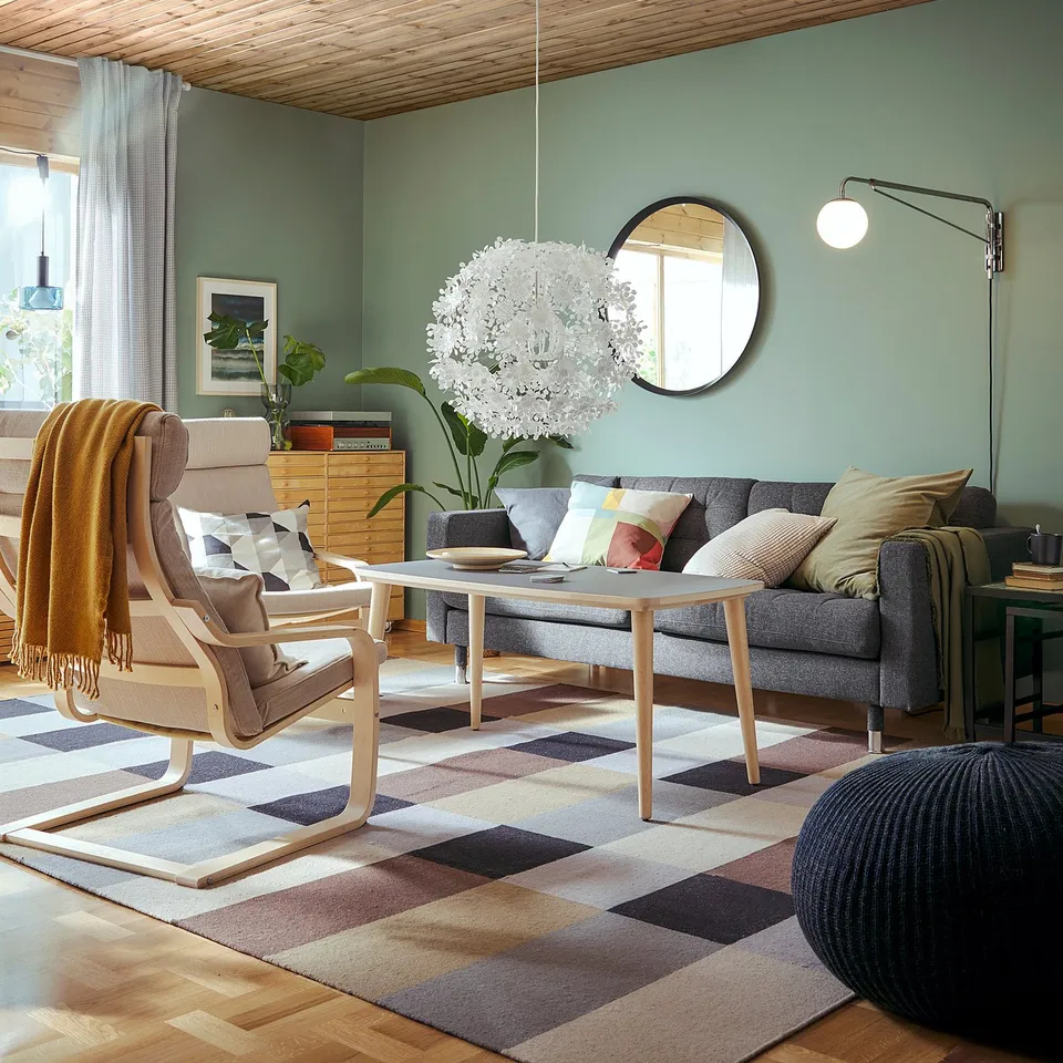 Modern living room with green walls, grey sofa, wooden ceiling, orange blanket chair, mirror, unique light, chequered rug, plants.