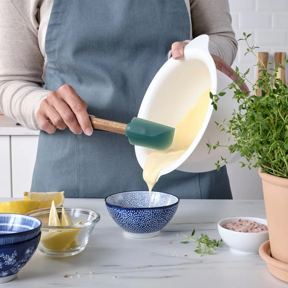 A person pours batter from a bowl using a green spatula. Various small bowls with ingredients are nearby. The spatula ensures no batter waste.
