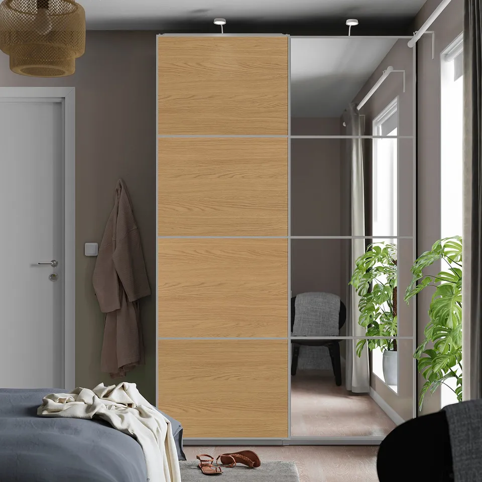 Modern bedroom with AULI closet, mirrored panels, soft close. Grey bed with brown sandals, large plant, sheer-curtained window.