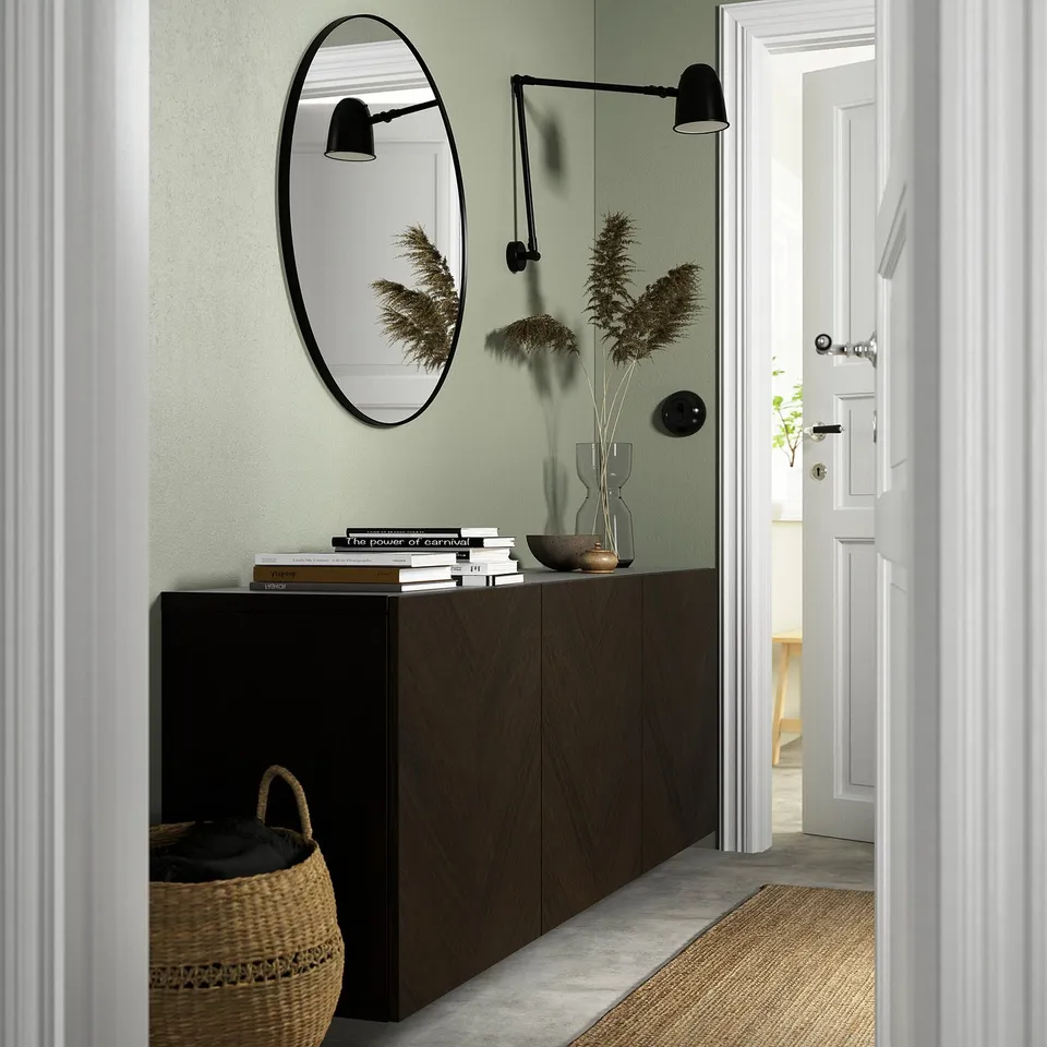 Modern entryway with BESTÅ sideboard, woven basket, light green wall, mirror, black sconce, dried plants in vase, and stack of books.