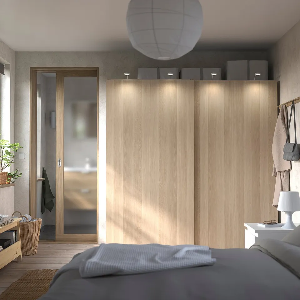 Modern bedroom light wood PAX wardrobe with sliding doors, grey bed, cosy lamp, and hanging accessories.