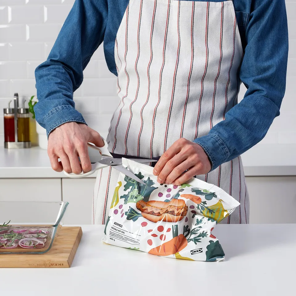 Person cutting open a bag with scissors while holding glass containers, wearing an apron in a kitchen.