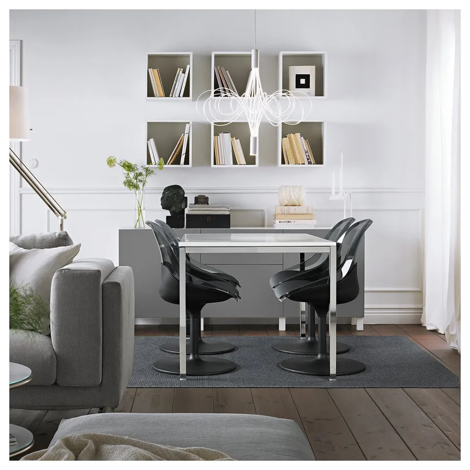 Modern dining area with glass-top table, BALTSAR chairs, and decor. Bright lighting and book-filled shelves enhance elegance.