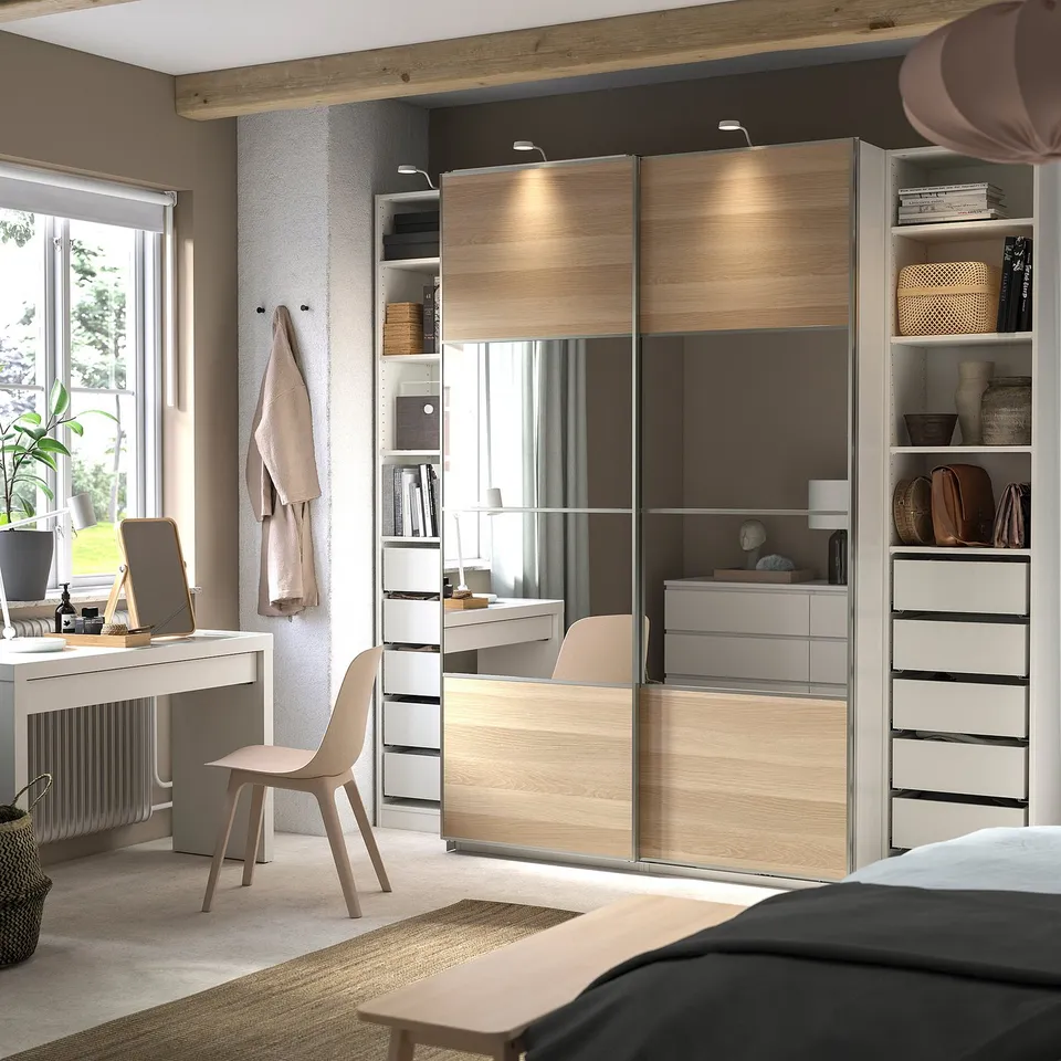 Modern bedroom with beige PAX wardrobe and sliding doors. Desk, chair, and shelves visible.