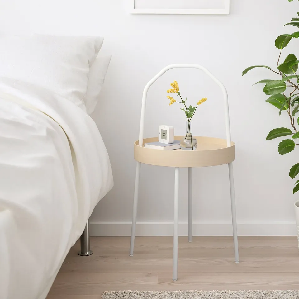 Small white BURVIK side table with wooden top, handle, and metal legs a yellow flower vase, alarm clock, and book beside a bed.