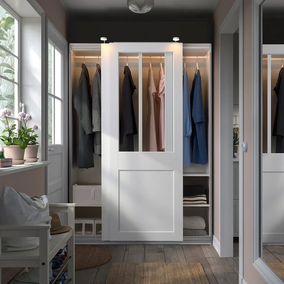 A white PAX wardrobe with glass doors displaying clothes. Its large, positioned between windows and a door, with a plant and basket nearby.