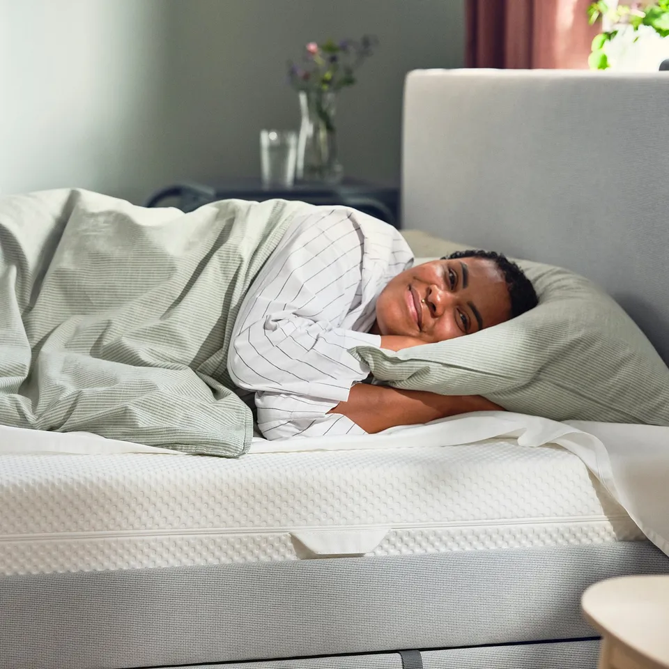 A person smiling on a white ÅBYGDA mattress, with a striped blanket over the person.