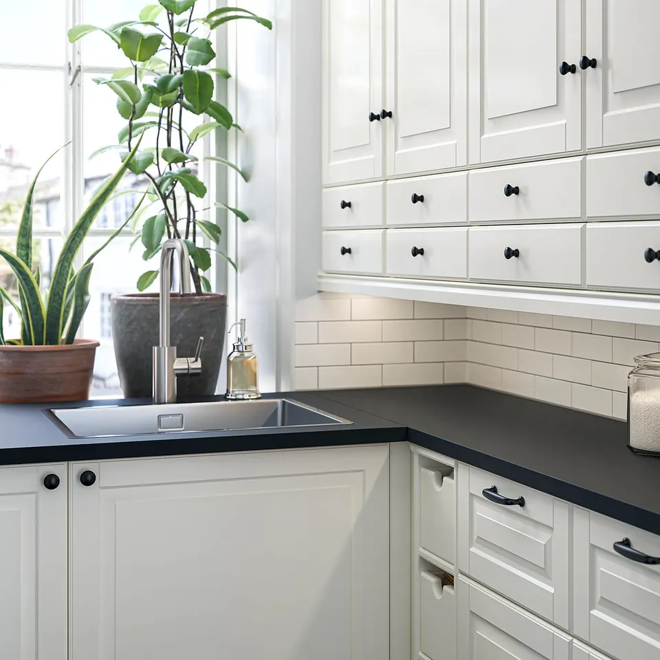 Modern white kitchen with black countertops, featuring sleek cabinets, black handles, and a potted plant near a window.