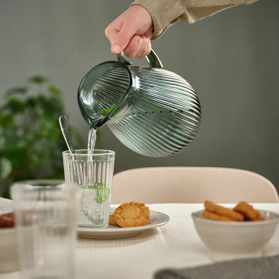 Person pouring water from a clear glass pitcher into a glass with cookies and snacks on a table.