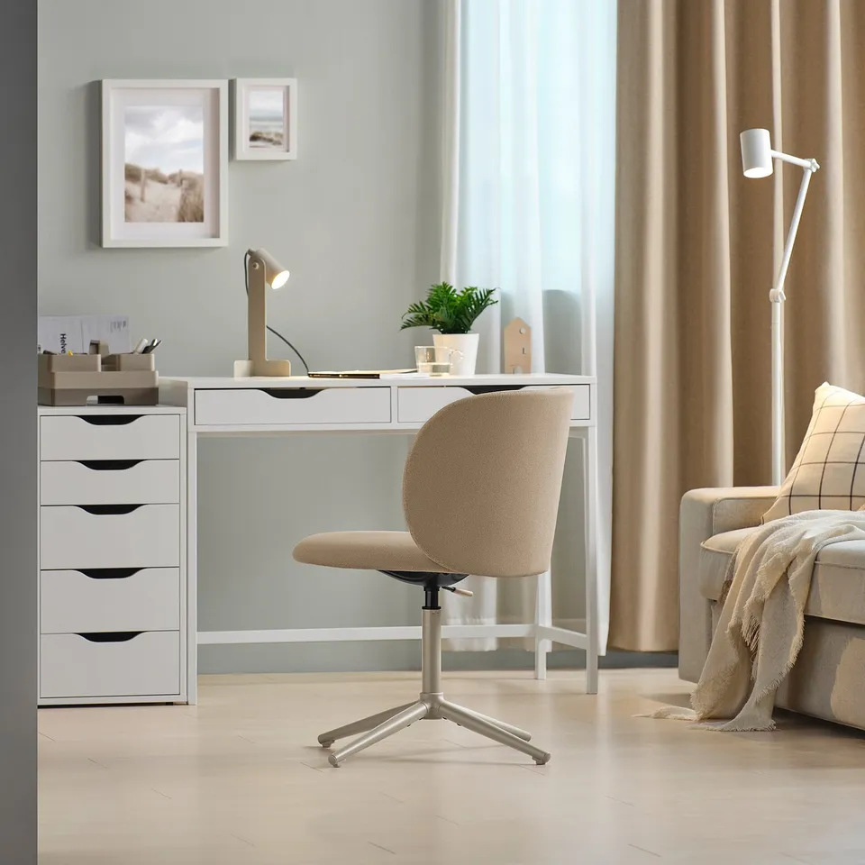 Modern home office with white KRYLBO swivel chair, white desk, drawer unit, framed pictures, beige sofa, and plant.