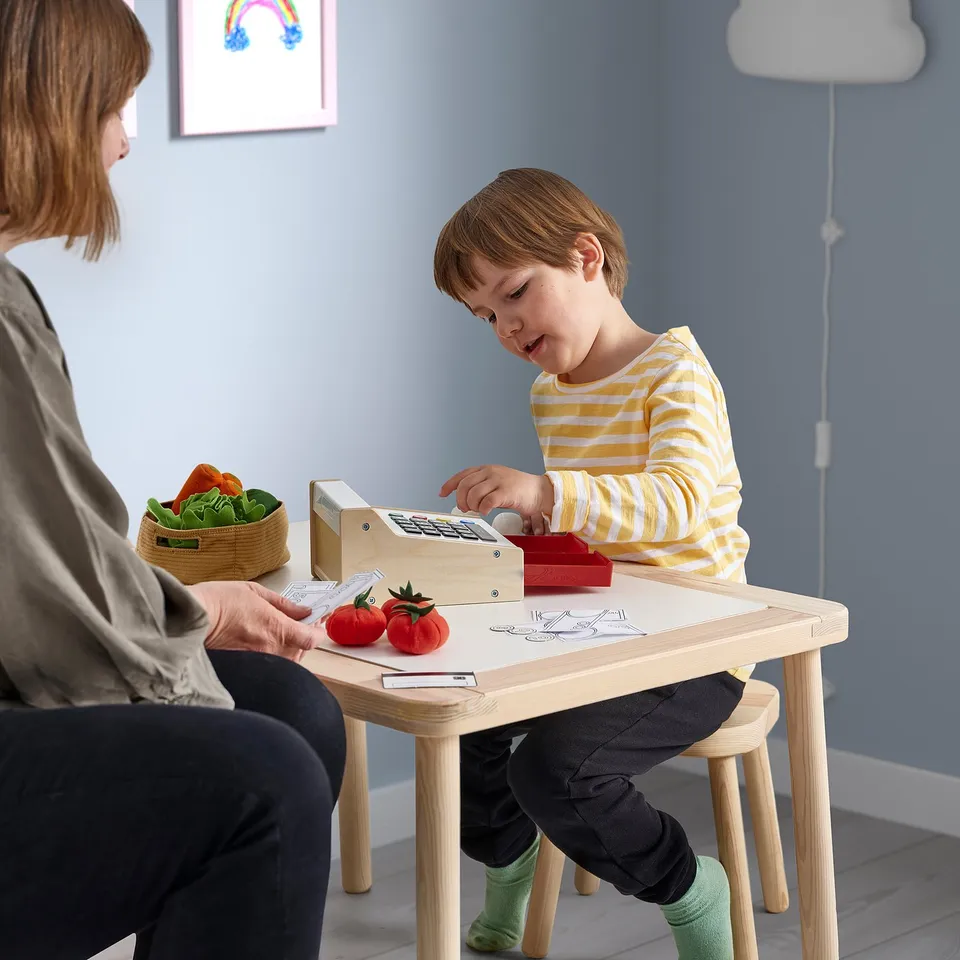 Child playing with DUKTIG cash register at wooden table. Person assists, tomatoes on table.