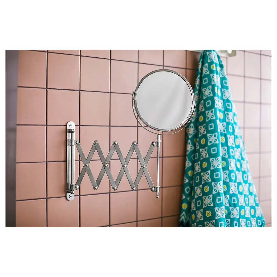 A FRÄCK magnifying mirror with silver frame, mounted on a pink-tiled wall, with a patterned cloth in the background.