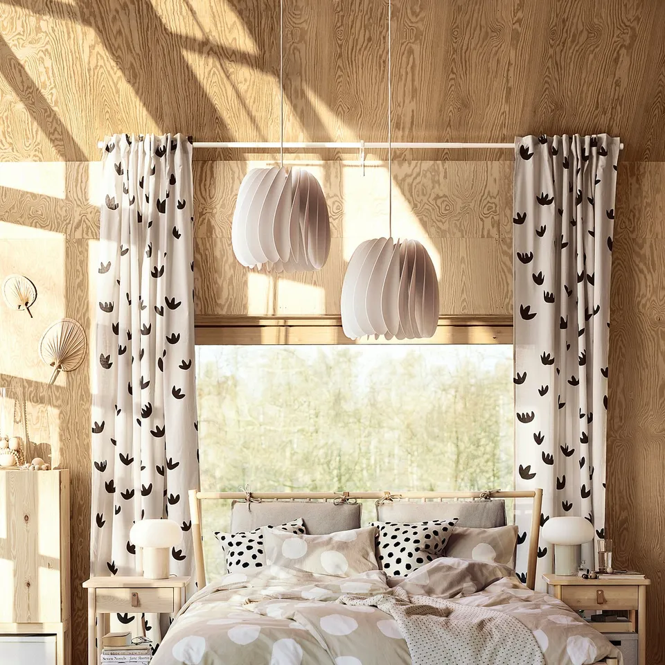 Cosy bedroom with light wooden decor, SKYMNINGEN white lamps, polka dot bedding, curtains with crowns, natural light through large window.