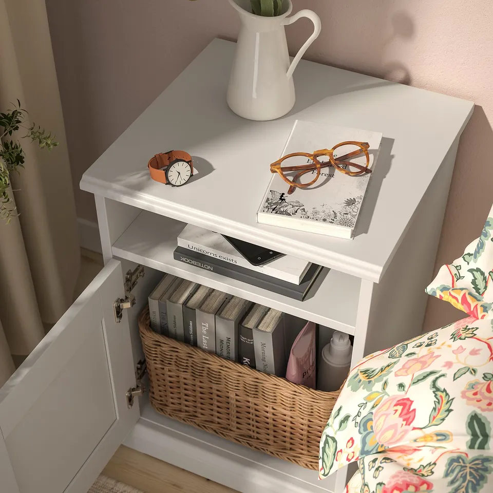 Bedside table with white surface, 2 shelves, books, basket, and supplies. Yellow watch, glasses, and white pitcher with flowers on top.