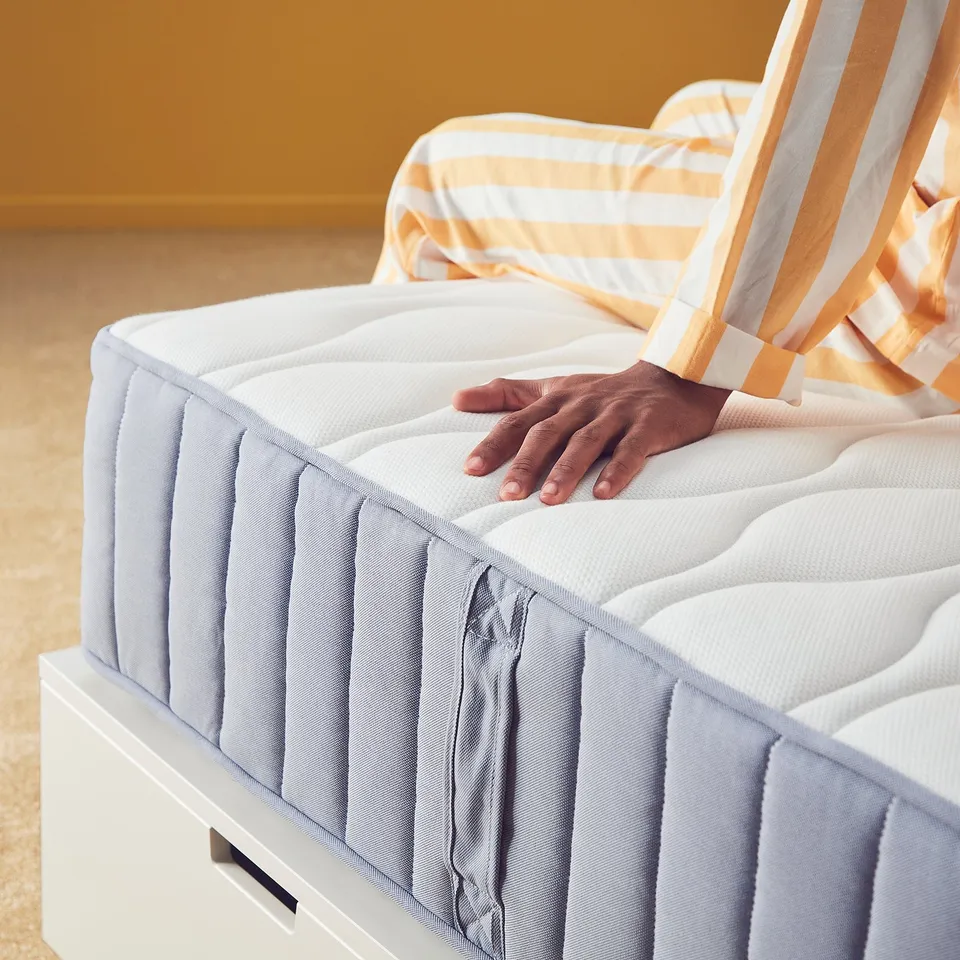 A person inspects a grey VÅGSTRANDA mattress, feeling the firm support.