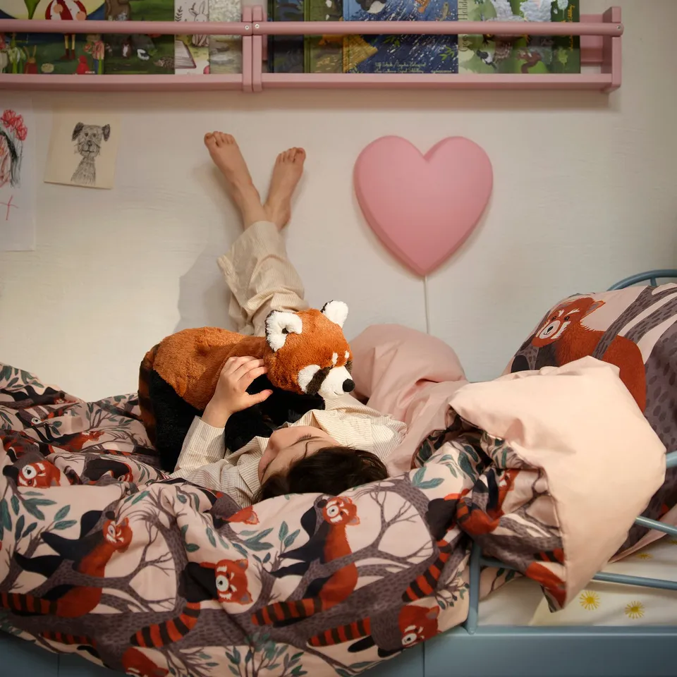 Child lying in bed, hugging a red panda stuffed animal, under a red panda-themed duvet. Bed has pink sheets and pillows.