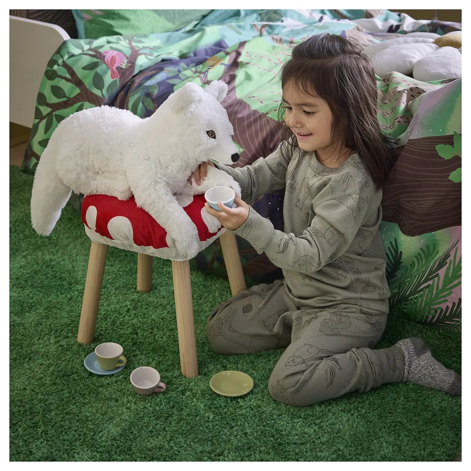 Young girl sits on green carpet, feeding a plush white fox toy from a tiny cup.