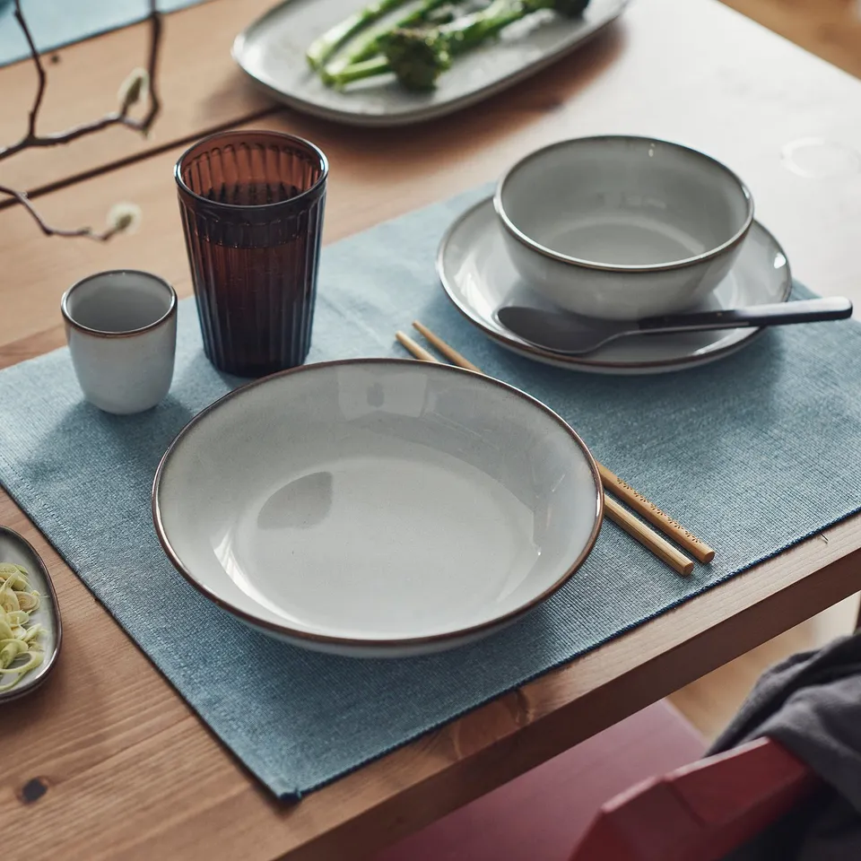 Table set with GLADELIG sandy-glazed deep plate, smaller than bowls, surrounded by chopsticks and cups on blue placemats.