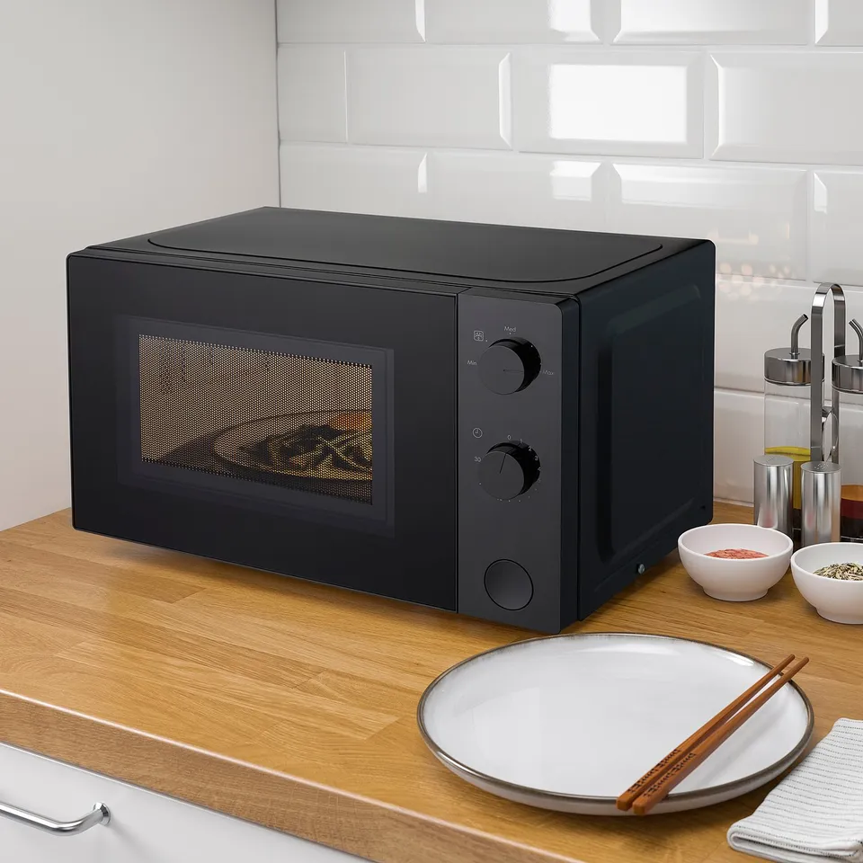 Black TILLREDA microwave on kitchen counter. Small, sleek design with black knobs. Placed near white dish, chopsticks, and spices.
