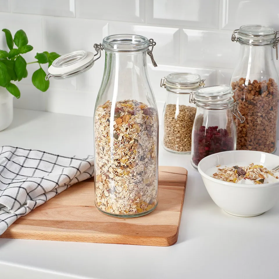 Clear glass jars, sealed with lids, store granola and dried foods on a wooden board. A bowl of granola sits nearby.