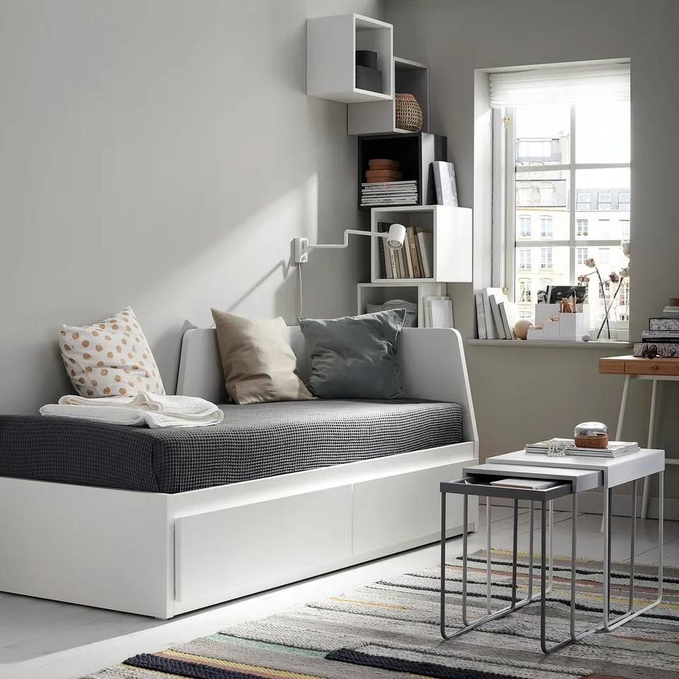 Modern minimalist living room with white FLEKKE sofa-bed, grey cushions, side tables, and bookshelf near a large window.