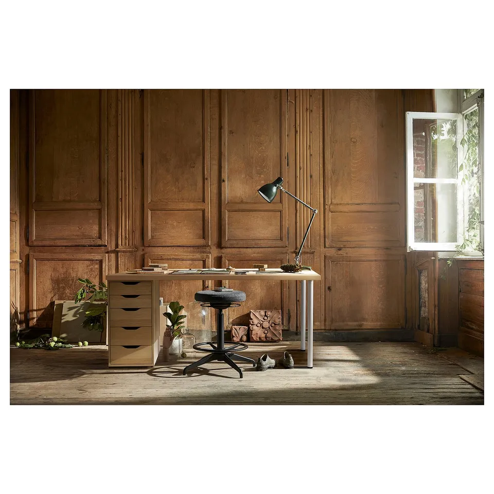 Desk setup with wooden panels, light-colored desk, black chair, and lamp.